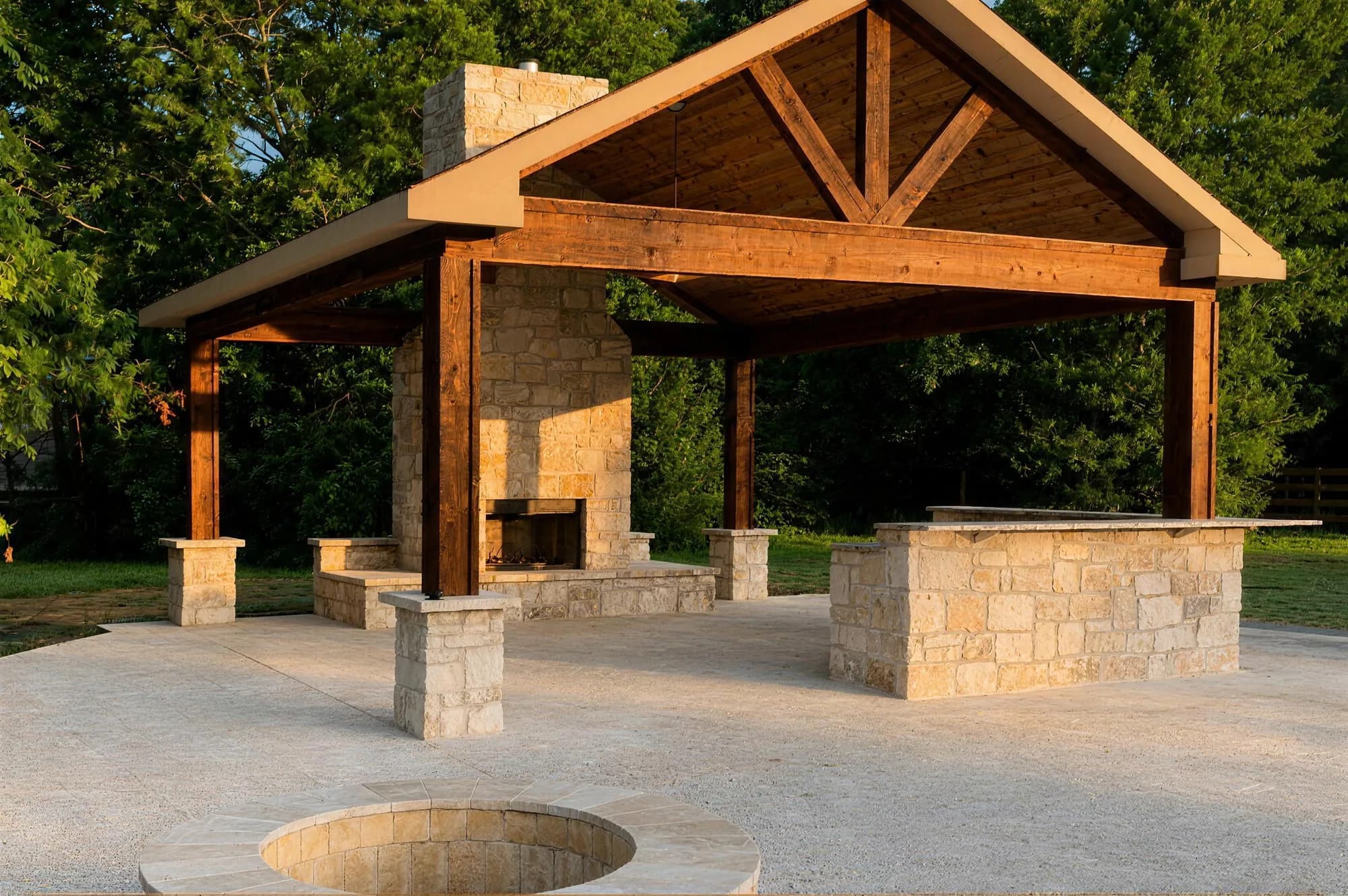 A gabled cedar pavilion with a stone fireplace and stone bar in golden afternoon light, on a stamped concrete patio with a round stone fire pit — a completed Royal Patio project in Prosper